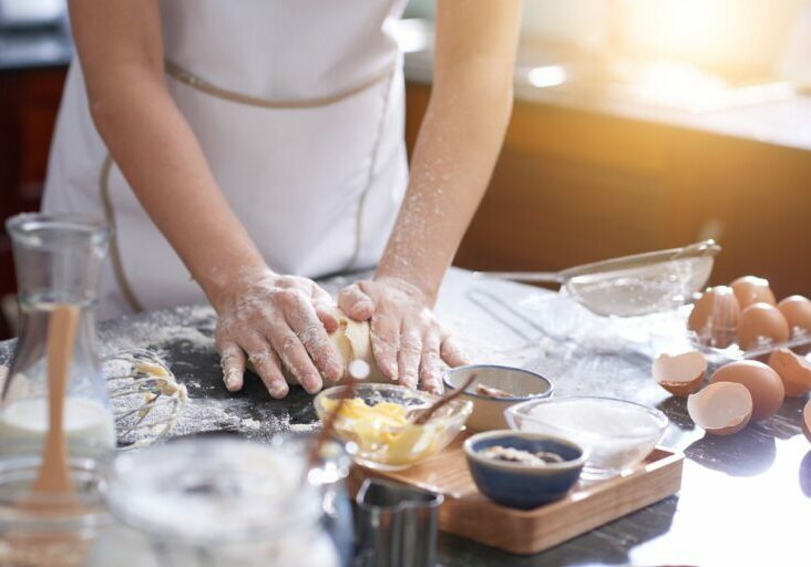 Hands of anonymous woman covered with flour kneading cookie dough on messy kitchen table, sunshine illuminating room