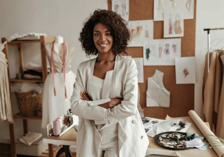 Beautiful curly brunette dark-skinned fashion designer poses in office, leans on table. Young lady in white suit crosses arms and smiles.